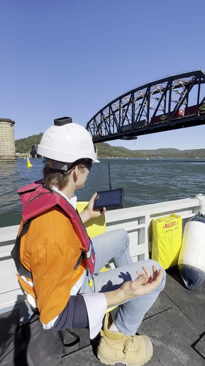 Worker in a life jacket and hard hat reading a handheld device on a boat beside a steel bridge