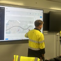 Two workers in high-visibility clothing reviewing a wall-mounted operations display