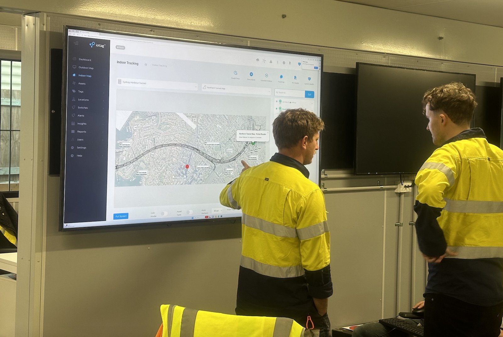 Two workers reviewing the iottag Atlas live tracking dashboard on a wall-mounted screen